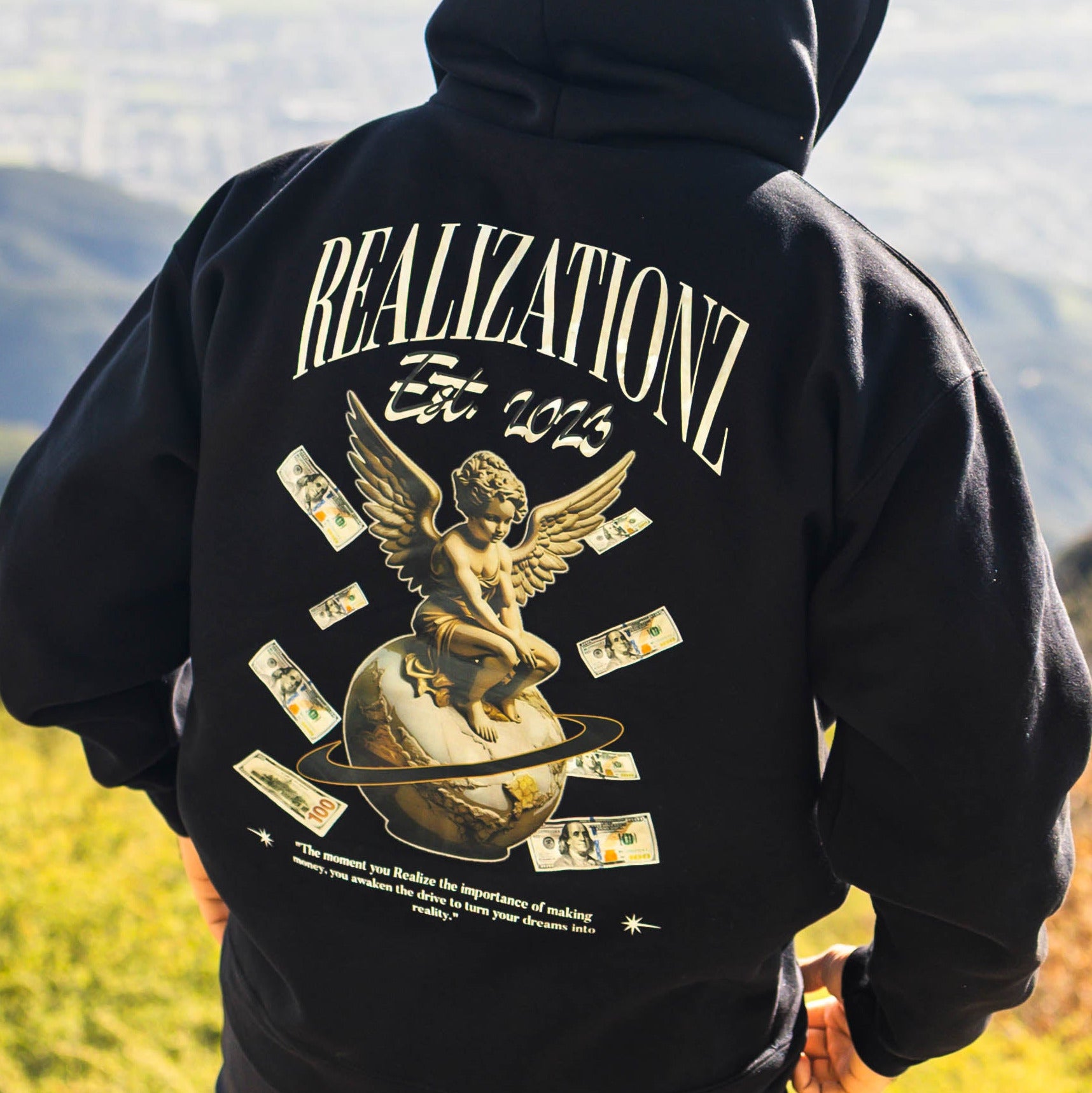 Person wearing a black hoodie with 'Realization' design, standing against a scenic background.